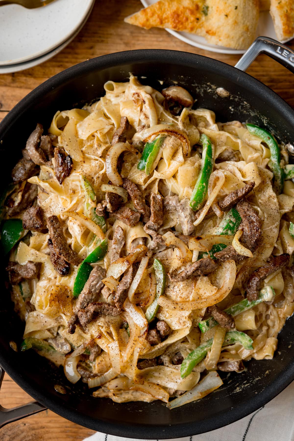 A tall, overhead image of Philly Cheesesteak Pasta. The pasta is in the centre of the image, in a black pan with a silver handle. To the right of the background, behind the pan, you can see a white and blue stripy napkin tucked behind the pan. In the top left of the background, you can slightly see the corner of two white bowls, stacked on top of each other. In the top right of the image, there is a white plate with a slice of garlic bread on top. This is all set on a wooden surface.