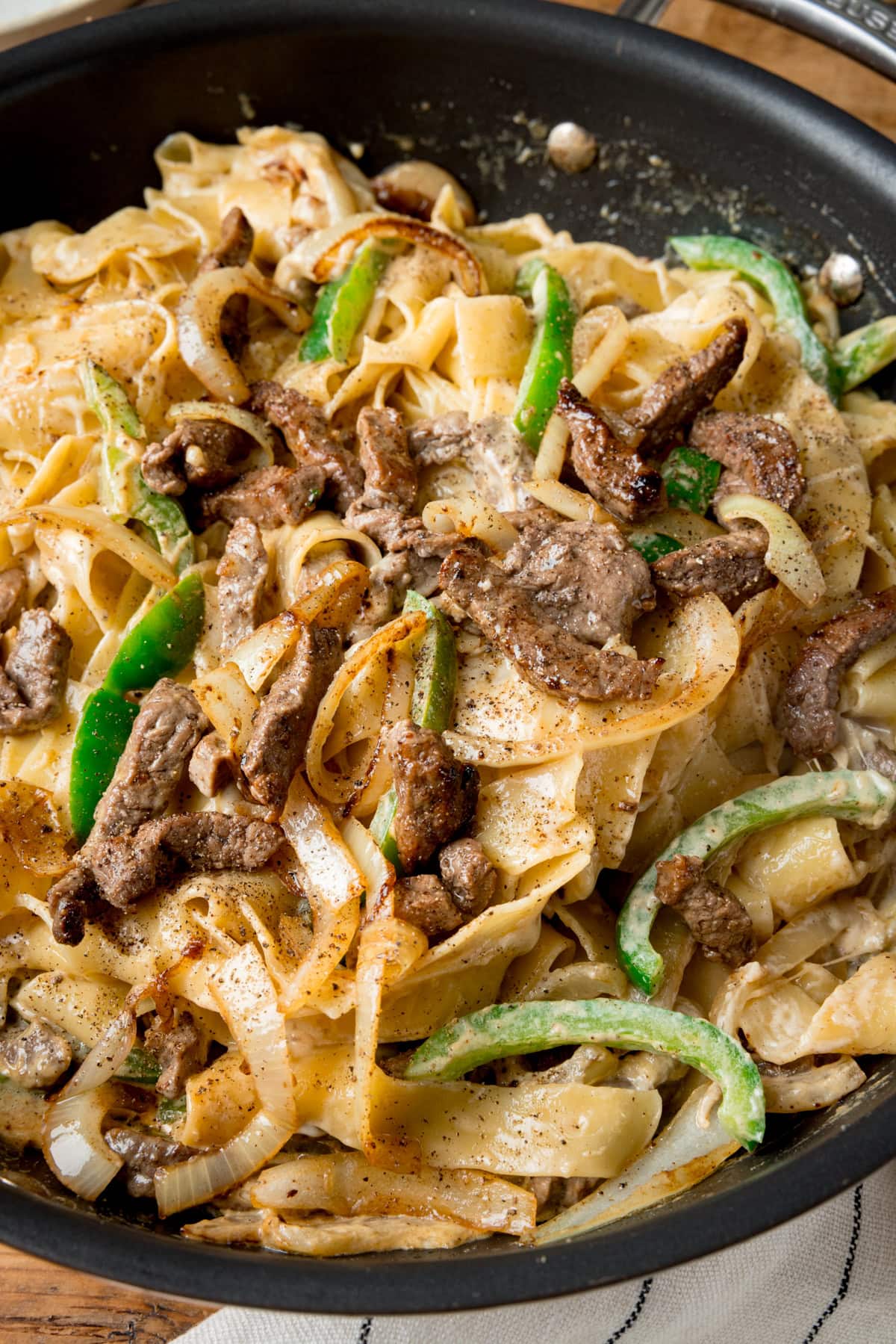 A tall image of Philly Cheesesteak Pasta. The pasta is in the centre of the image, in a black pan with a silver handle. To the right of the background, behind the pan, you can see a white and blue stripy napkin tucked behind the pan. This is all set on a wooden surface.