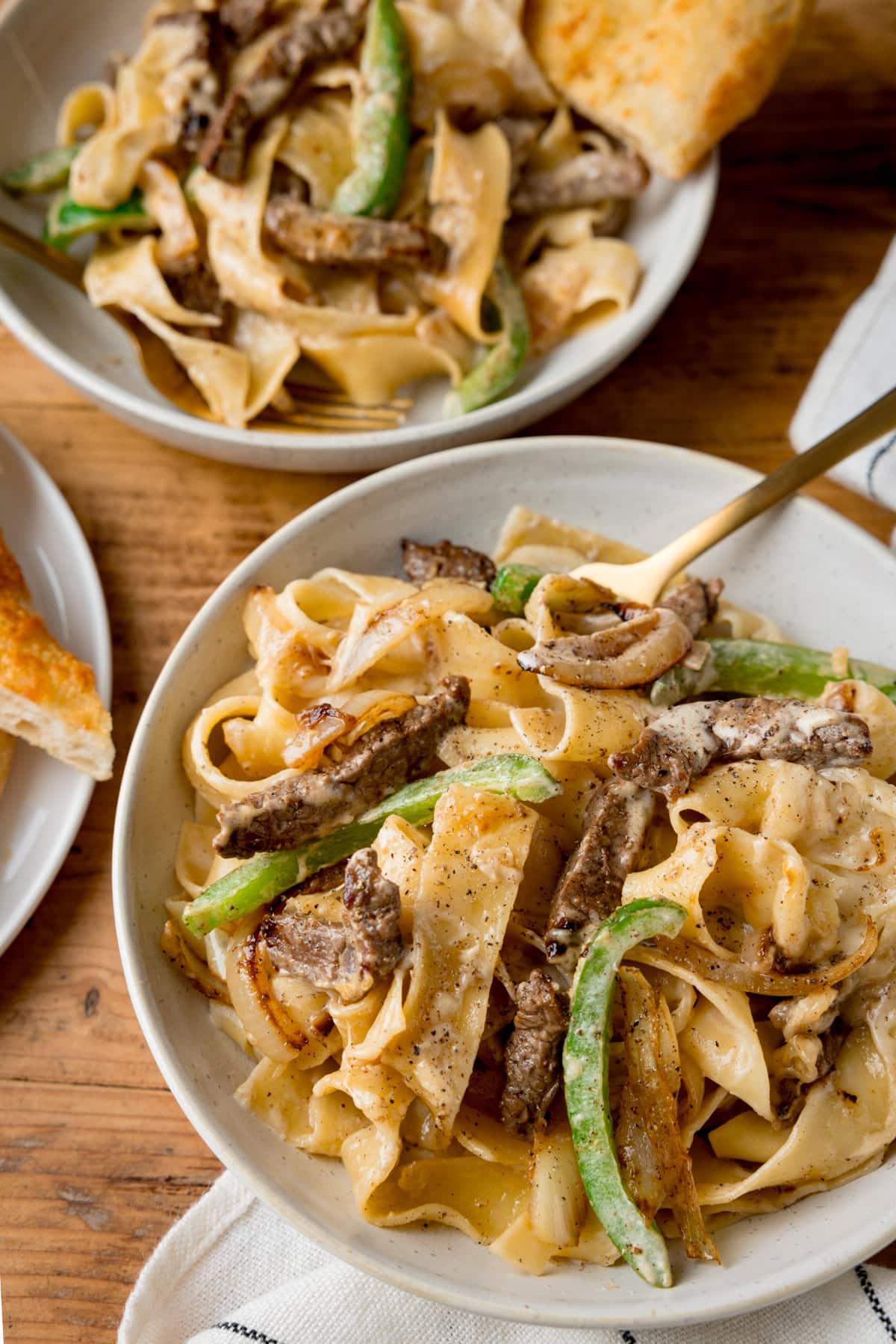 A tall, overhead shot of Philly cheesesteak pasta. The pasta is in a white bowl, in the centre of the image. There is a gold fork sticking out of the dish. At the bottom of the image, there is a white and blue striped napkin tucked beneath the bowl. At the top of the image, there is a second white bowl of pasta, with a slice of garlic bread on top of the pasta. At the left of the image, you can slightly see a white plate with more garlic bread on it. This is all set on a wooden surface.