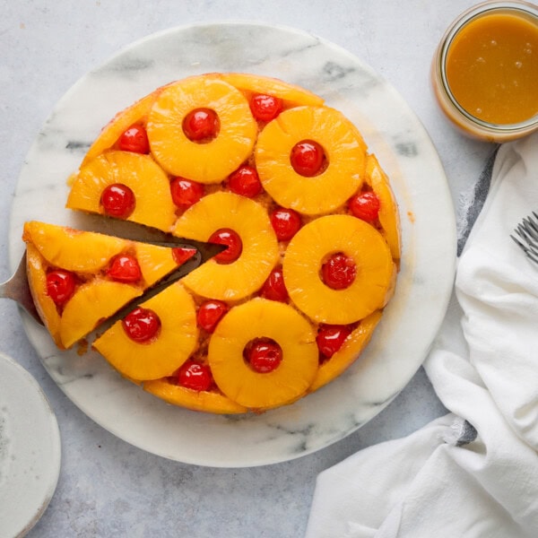 A tall, overhead shot of pineapple upside down cake on a white marble cake stand. A slice of the cake has been cut from the cake and you can see a silver cake slice tucked underneath the separated slice. On the top right of the image, there is a silver spoon, next to it is a clear jar of pineapple syrup. To the left of the image, you can slightly see a white plate. To the right of the image, there is a white napkin tucked underneath the main cake stand. This is all set on a white surface.