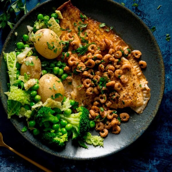 A square shot of a bowl of Plaice with Brown Butter Shrimp served on a black dish with peas, cabbage, broccoli and new potatoes served on the side. Beneath the main dish, there is a navy blue napkin with a gold fork on top, which is on the bottom left of the image. In the background, there are small parsley leaves sprinkled around. This is set on a dark blue background.