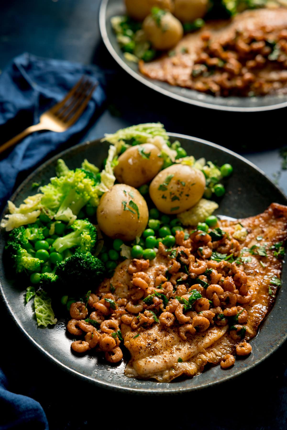 A tall, closeup shot of a bowl of Plaice with Brown Butter Shrimp served on a black dish with peas, cabbage, broccoli and new potatoes served on the side. There is a second identical dish of Plaice with Brown Butter Shrimp on the left of the background. Beneath the second dish, there is a navy blue napkin with a gold fork on top. In the background, there are small parsley leaves sprinkled around. This is set on a dark blue background.