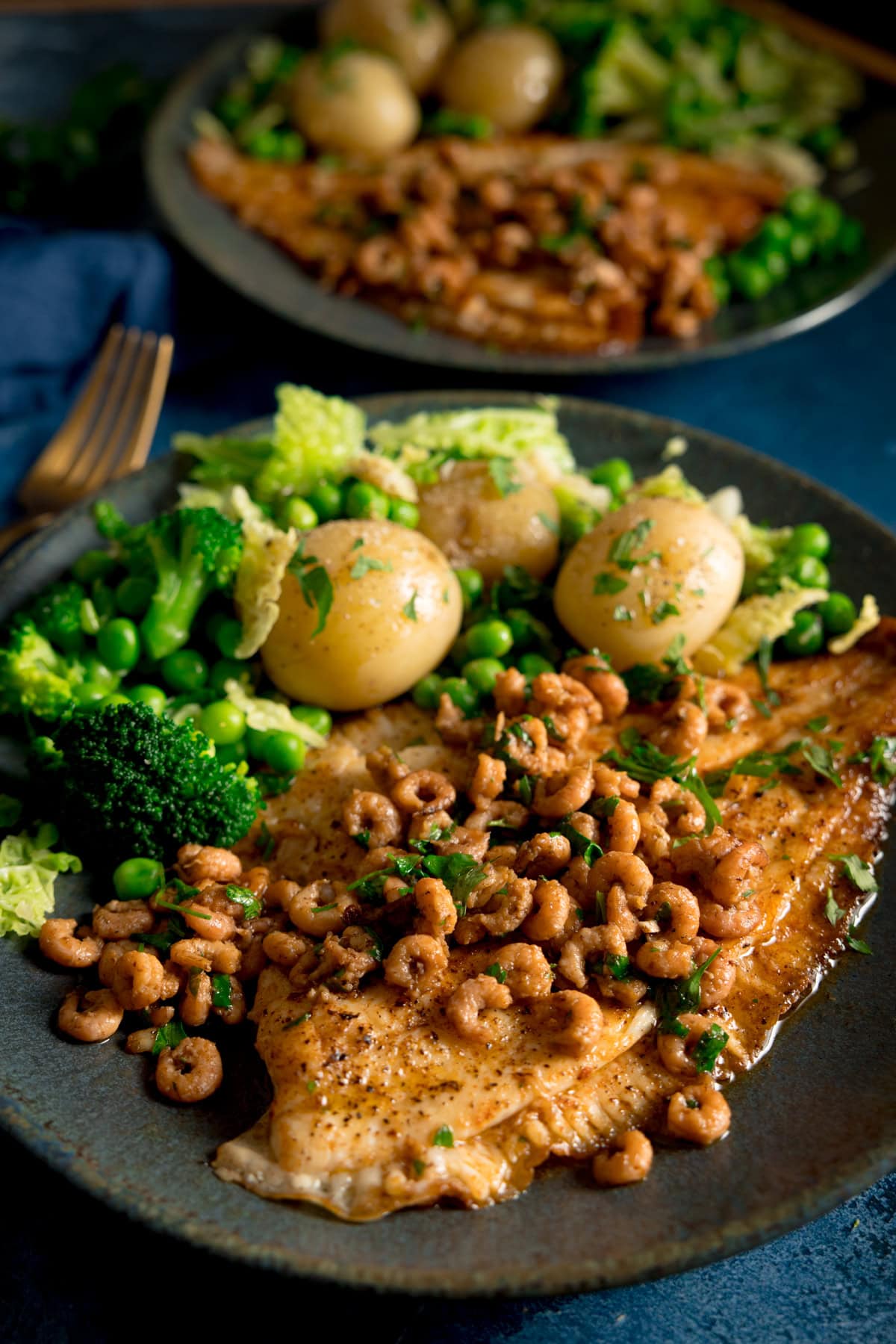 A tall shot of a bowl of Plaice with Brown Butter Shrimp served on a black dish with peas, cabbage, broccoli and new potatoes served on the side. There is a second identical dish of Plaice with Brown Butter Shrimp on the left of the background. Beneath the second dish, there is a navy blue napkin with a gold fork on top. In the background, there are small parsley leaves sprinkled around. This is set on a dark blue background.