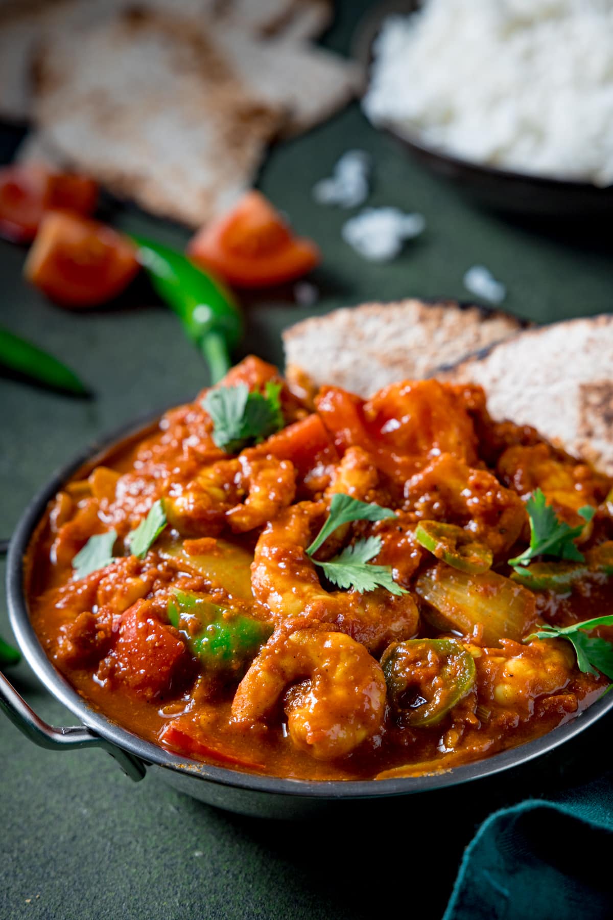 Picture of a prawn curry in a balti dish with a bowl of boiled rice and some chapati in the background.