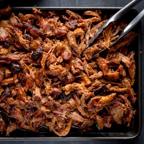 A square, overhead image of BBQ Pulled Pork. The pulled pork is in a rectangular black pan in the centre of the image. In the top right of the pan, there is a pair of tongs sticking out of the pork at an angle. This is all set on a grey background.