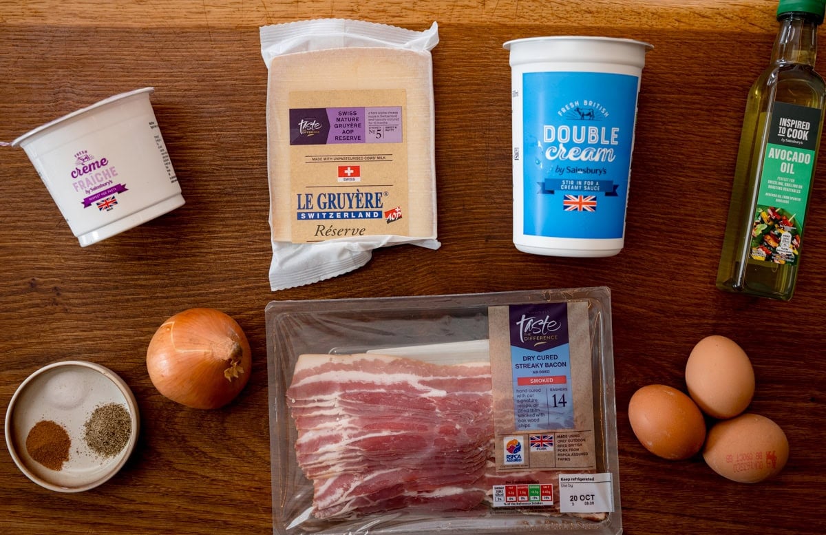 A wide overhead shot of the ingredients for the filling of Quiche Lorraine. The ingredients are laid out on a wooden cutting board. The ingredients are as follows: Creme Fraiche, onion, double cream, Gruyere, oil, eggs, smoked bacon, salt, black pepper, and nutmeg.