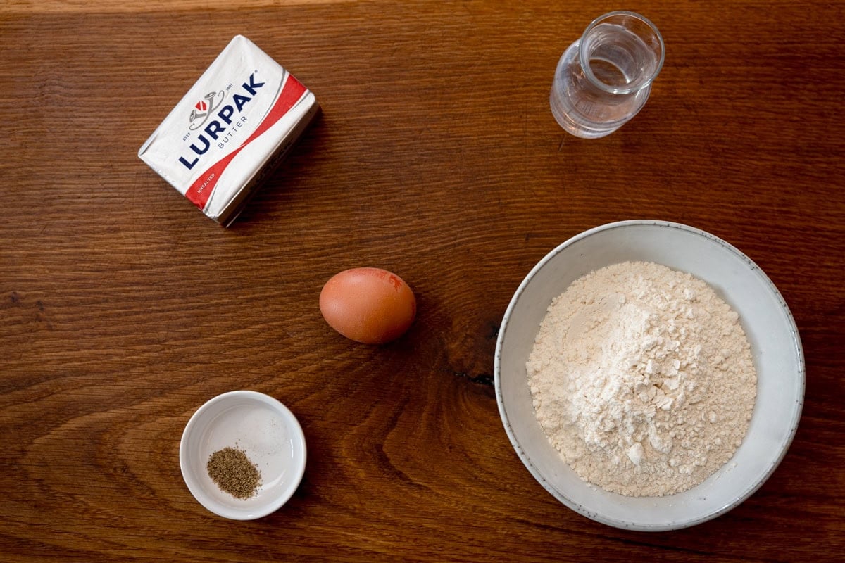 A wide, overhead shot of the ingredients for the pastry of Quiche Lorraine. The ingredients are laid out on a wooden cutting board. They are as follows: flour, butter, egg, water, salt and pepper.