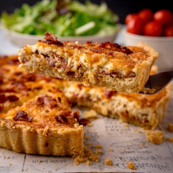 A square image of Quiche Lorraine. The quiche is in the centre of the image, a slice of the quiche has been removed and is being shown in the foreground. The slice is on top of a black, plastic spatula, which is coming from the right side of the image. In the background, you can see one large white bowl filled with salad, and to the right of that, a smaller white bowl filled with cherry tomatoes. This is all set on a white newspaper baking parchment surface, and a black background.