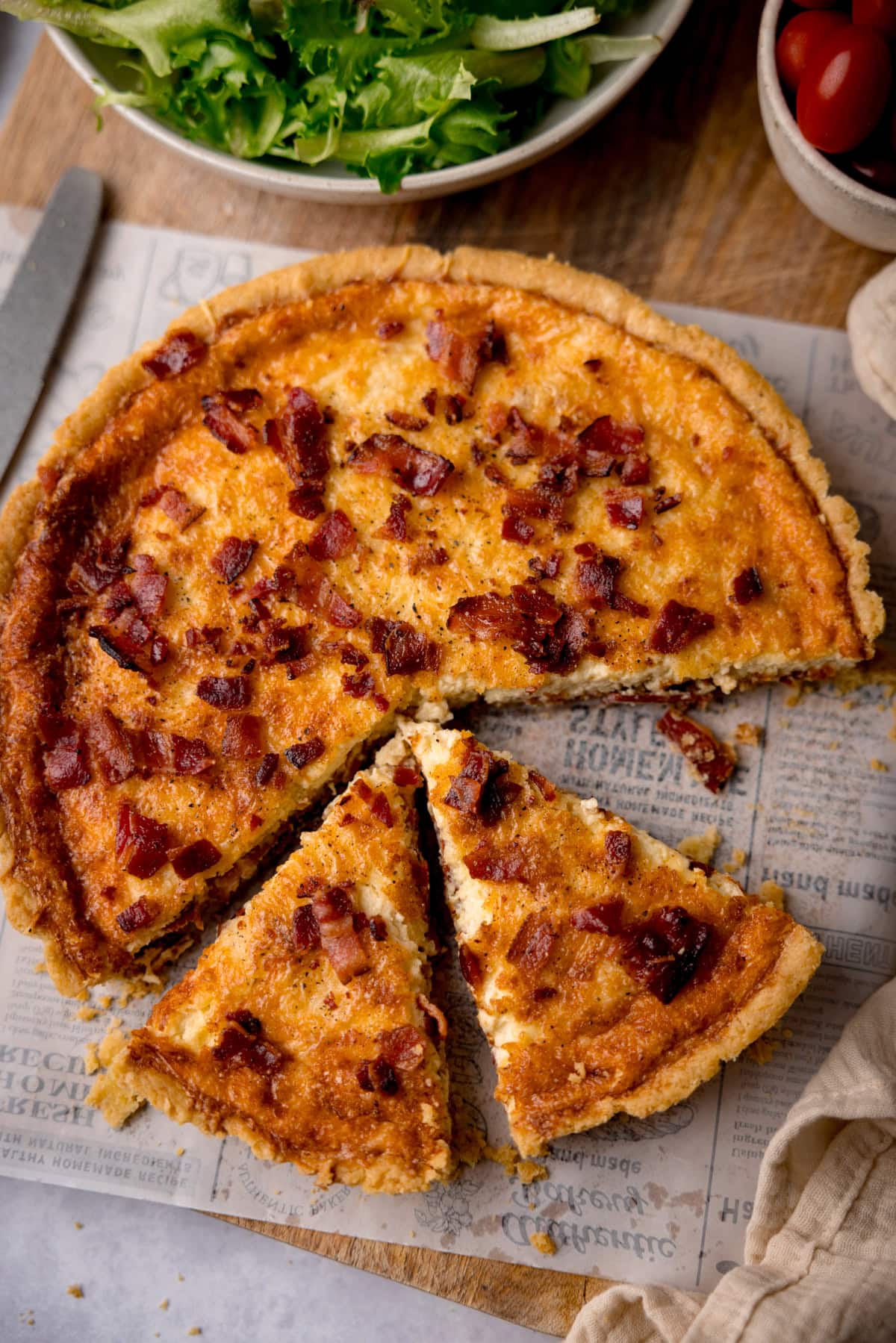 A tall, overhead shot of Quiche Lorraine. The quiche is in the centre of the image, placed on top of a piece off-white newspaper baking parchment. The quiche is whole, apart from three slices taken out of it, two of the slices are still in place, the other piece is missing. To the left of the quiche, there is a silver spatula. To the bottom right of the quiche, there is an off-white napkin. At the top of the image, there is a large white bowl filled with salad, and to the right of that, there is a smaller white bowl filled with cherry tomatoes. This is all on top of a wooden cutting board, which is on top of a white surface.