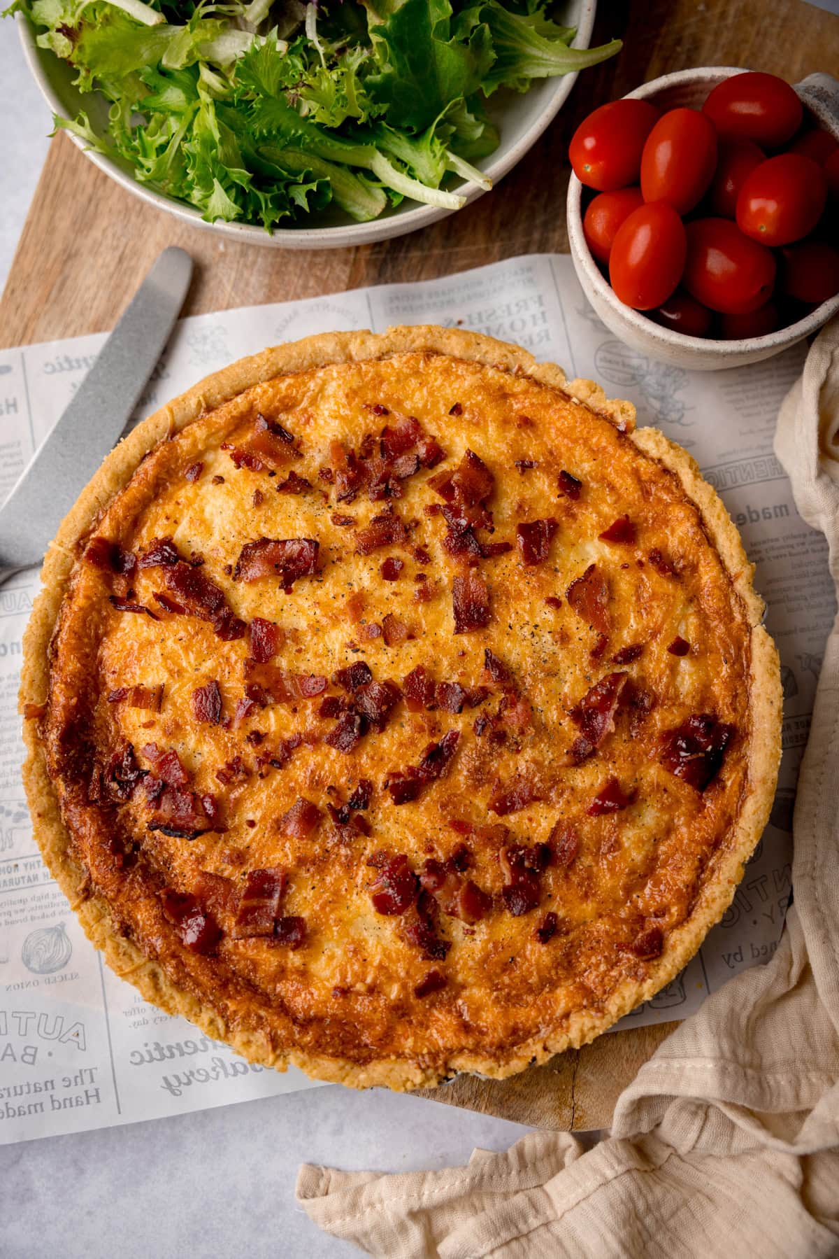 A tall, overhead shot of Quiche Lorraine. The quiche is in the centre of the image, placed on top of a piece of white newspaper baking parchment. To the left of the quiche, there is a silver spatula. To the bottom right of the quiche, there is an off-white napkin. In the top of the image, there is a large white bowl filled with salad, and to the right of that, there is a smaller white bowl filled with cherry tomatoes. This is all on top of a wooden cutting board, which is on top of a white surface.