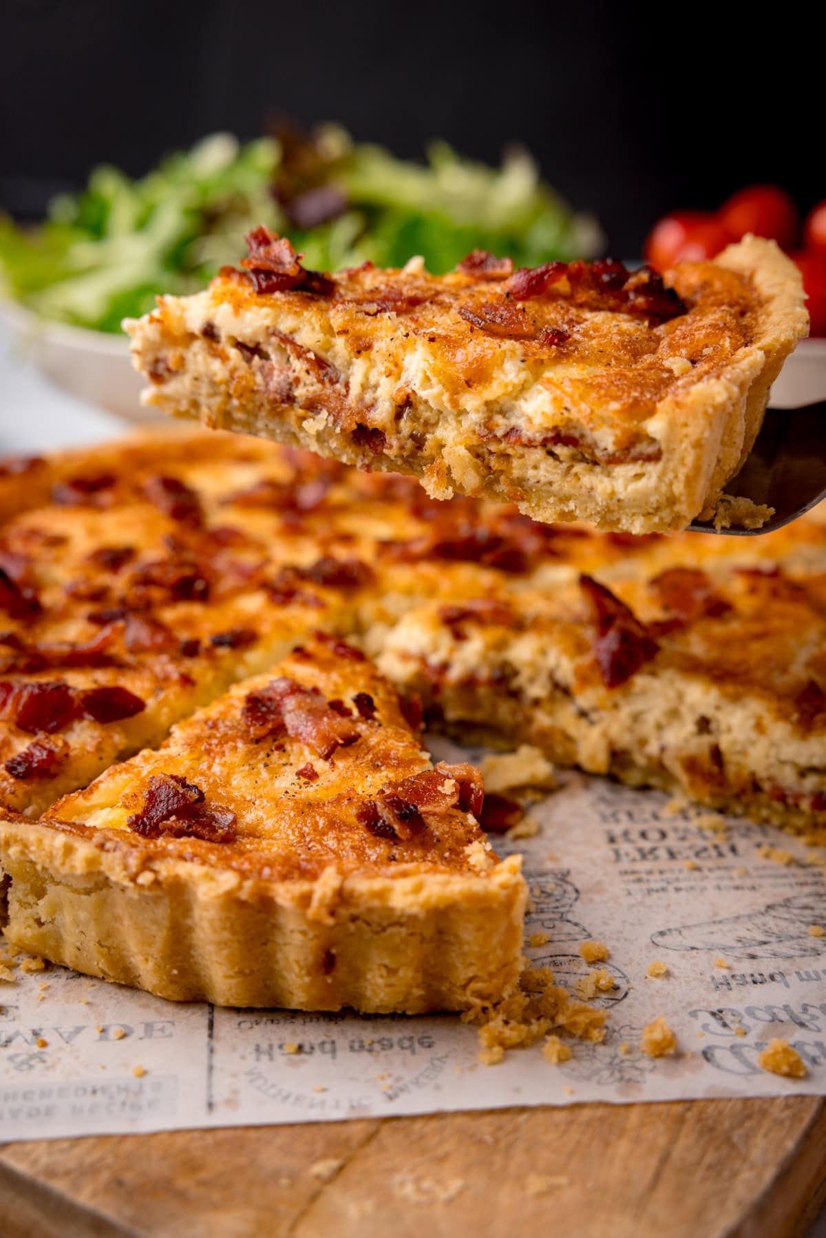 A tall image of Quiche Lorraine. The quiche is in the centre of the image, a slice of the quiche has been removed and is being shown in the foreground. The slice is on top of a black, plastic spatula, which is coming from the right side of the image. In the background, you can see one large white bowl filled with salad, and to the right of that, a smaller white bowl filled with cherry tomatoes. This is all set on a white newspaper baking parchment piece, which is on top of a wooden cutting board, and a black background.