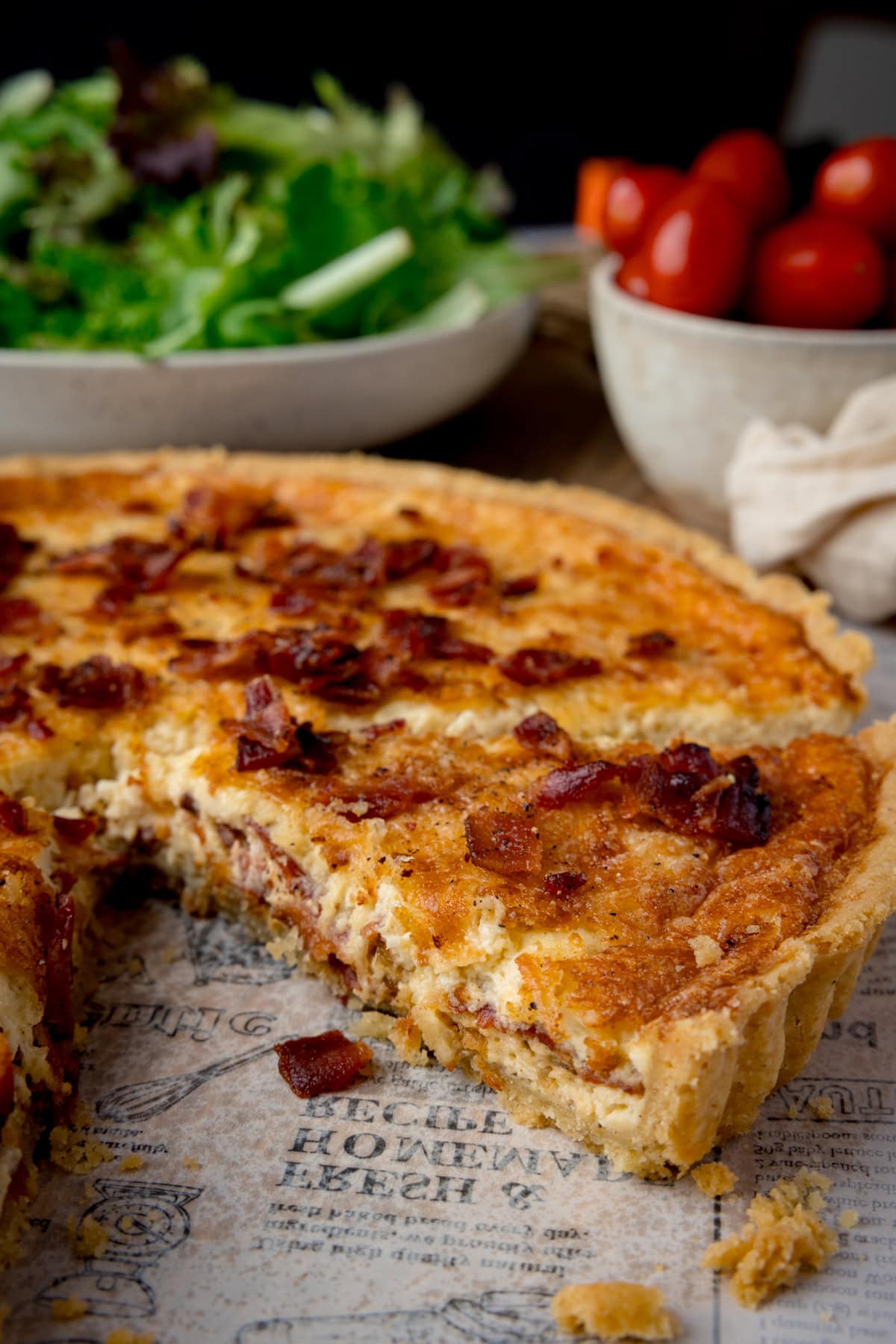 A tall image of Quiche Lorraine. The quiche is in the centre of the image, and one slice of the quiche is visibly missing. In the background, you can see one large white bowl filled with salad, and to the right of that, a smaller white bowl filled with cherry tomatoes. This is all set on a white newspaper baking parchment piece, which is on top of a wooden cutting board, and a black background.