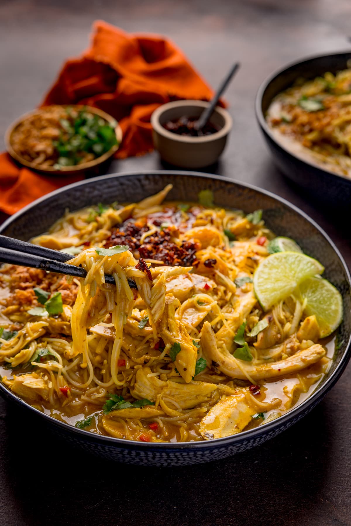 Close up picture of some black chopsticks picking up some chicken and noodles from a bowl full of chicken laksa soup.