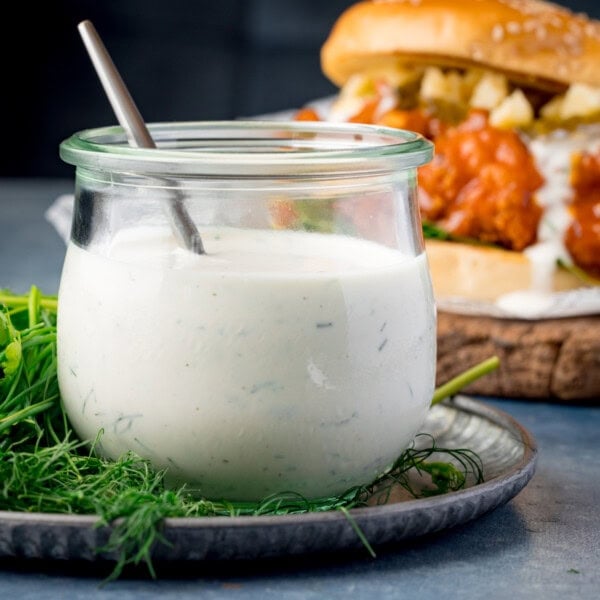 A square shot of Homemade Ranch Dressing. The dressing is in a clear glass jar in the centre of the image, there is a silver spoon sticking out of the jar with the dressing. The jar is placed on top of a black plate which has dill and coriander sprigs on it, surrounding the jar. On the right side of the background, there is a buffalo chicken burger (with the homemade ranch on it), placed on a wooden cutting board. This is all set on a light blue surface, with a dark blue background.