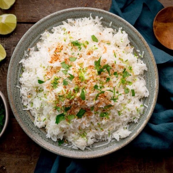 A square, overhead image of Rice Cooker Coconut Rice. The rice is in a large, grey bowl in the centre of the image and is topped with toasted coconut, coriander and lime zest. On the right side of the image, there is a dark teal napkin tucked underneath the bowl. On top of the napkin, in the right corner of the image, you can slightly see a large wooden spoon. In the top left corner of the image, you can see three lime wedges on the surface. Below that you can slightly see a light grey dish, filled with coriander. This is all set on a dark wooden background.