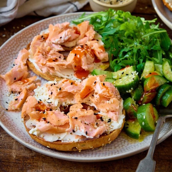 A square shot of Salmon Cream Cheese Bagels served on a light grey plate. There is a silver fork resting on the bottom of the plate. In the top left of the image, there is a white napkin with small grey stripes, a light brown dish with mixed sesame seeds in it. And in the top right of the image, you can slightly see the corner of a second identical plate of Salmon Cream Cheese Bagels. This is all set on a wooden background.