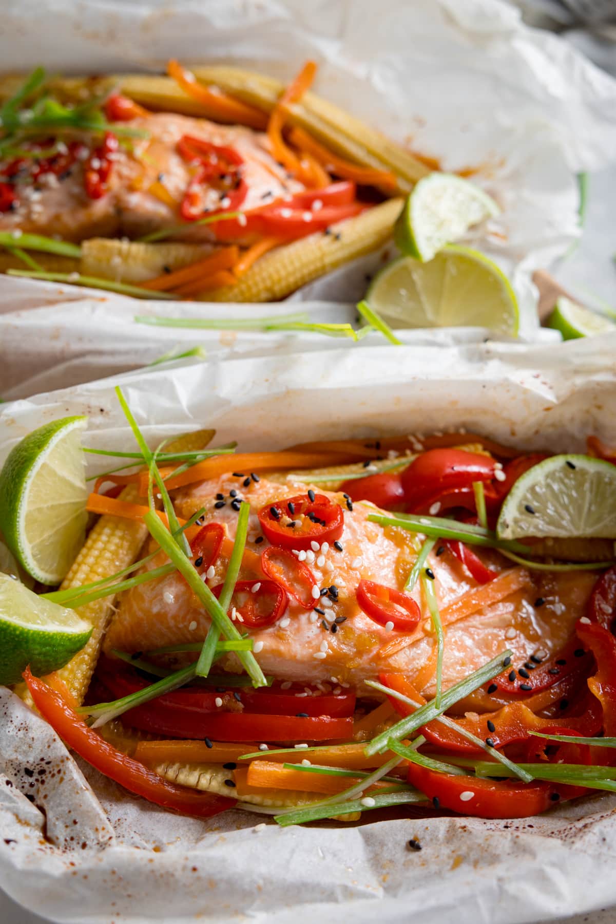 Close up overhead image of two salmon fillets cooked in individual baking parchment parcels with slices of chilli, peppers, babycorn, carrot and spring onion. Also topped with wedges of lime. The parcels are open.