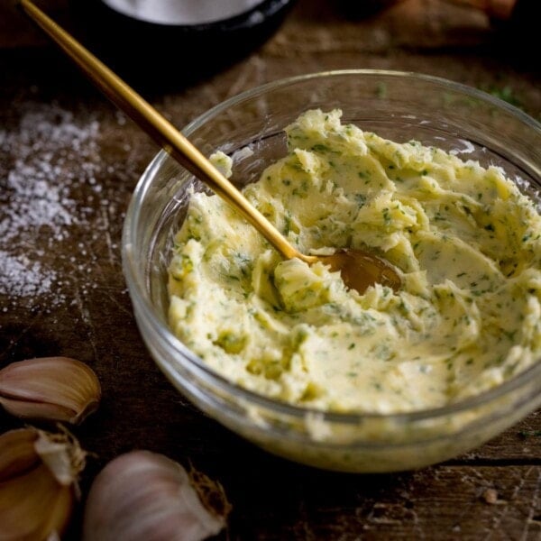 A bowl of garlic butter with a wooden spoon in it, on a wooden table with salt and bulbs of garlic on the side of the bowl