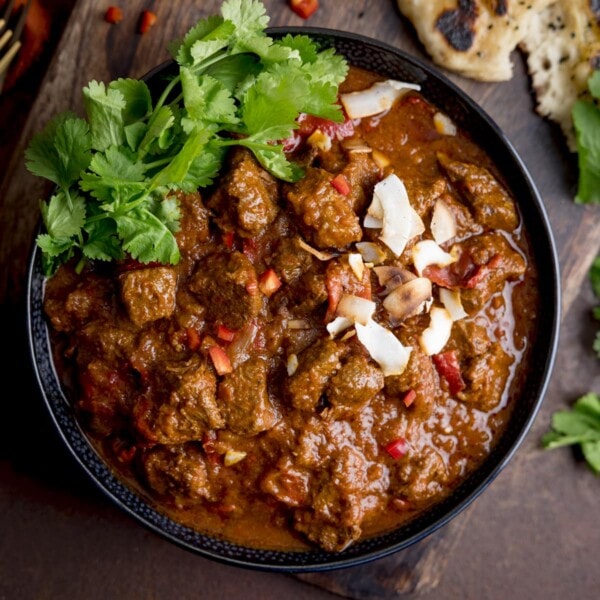 Square image of beef curry, topped with coriander, chillies and toasted coconut on a black bowl. The bowl is on a dark brown surface and there are some ingredients scattered around the bowl, just in shot.