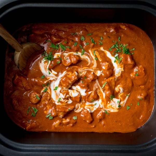 A square, overhead shot of Slow Cooker Chicken Curry in a rectangular, black slow cooker. The curry is topped with chopped coriander and there is a wooden spoon in the top left side of the dish.