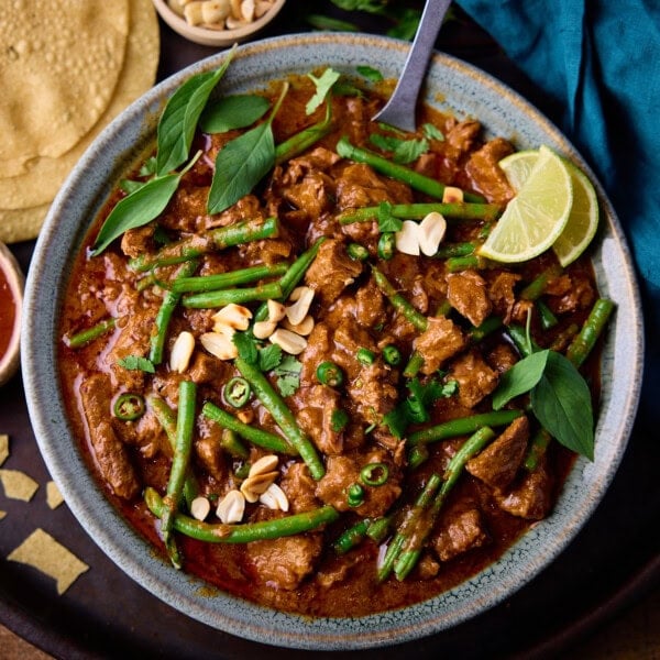 A square, overhead shot of Slow Cooker Thai Red Beef Curry in a light blue bowl. There is a silver spoon sticking out of the dish. To the left of the bowl, you can slightly see a small white dish filled with sweet chilli sauce, above that you can see a stack of popadoms. Directly above the main bowl, you can see a small white dish filled with peanuts. In the top right corner of the image, there is a blue napkin tucked beside the bowl. All of this is on top of a black tray, which is on top of a wooden surface.