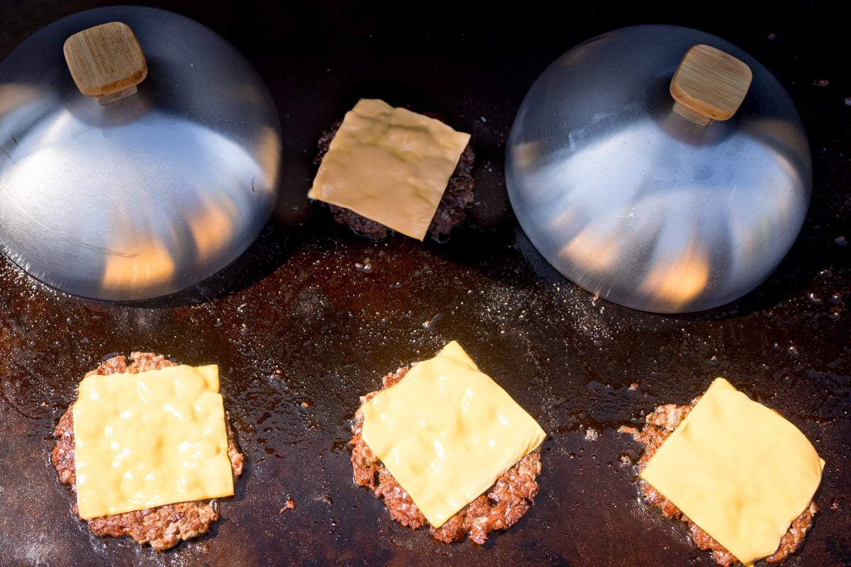 A wide, overhead shot of smash burgers cooking on a flat-top BBQ. There are 6 burgers on the BBQ, all with a square slice of cheese on them. The top left and top right burgers both have silver burger domes, with wooden handles covering them.