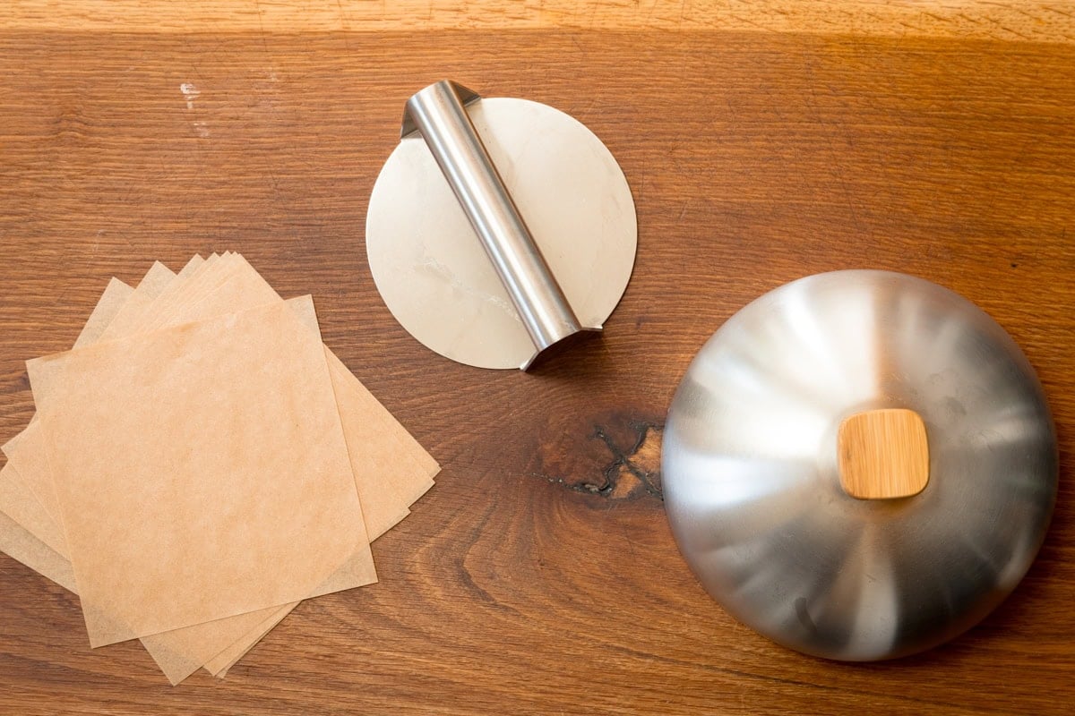 A wide, overhead shot of the equipment needed for smash burgers. There are a few squares of brown parchment paper on the left side of the surface, next to it, there is a silver burger press, and next to that, there is a silver burger dome with a wooden handle. This is all set on a wooden surface.