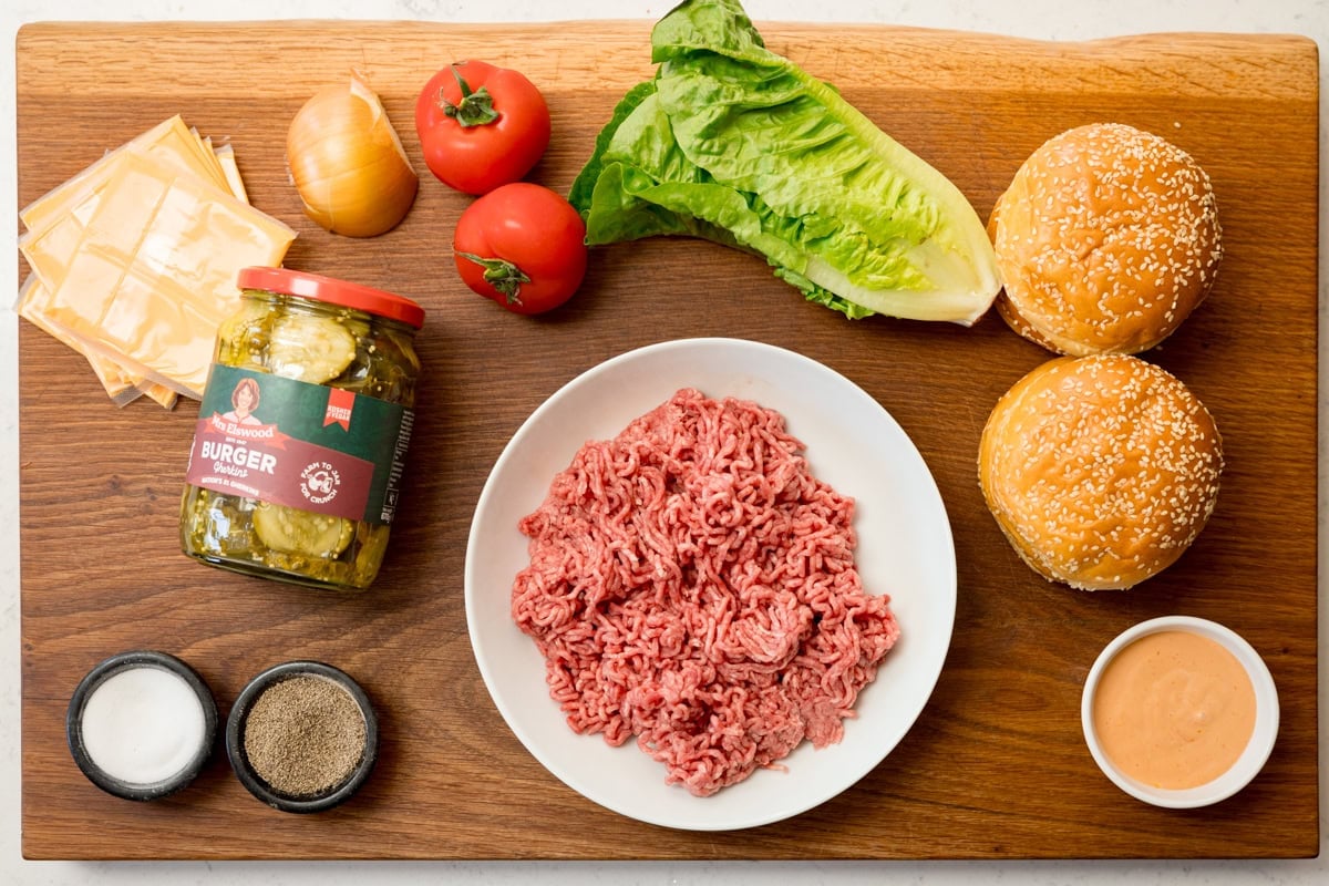 A wide, overhead shot of the ingredients for smash burgers. They are laid out on a wooden board. They are as follows: minced beef, salt, pepper, burger cheese slices, brioche buns, lettuce, tomatoes, burger sauce, onion, and pickled gherkins. This is all set on a wooden board.