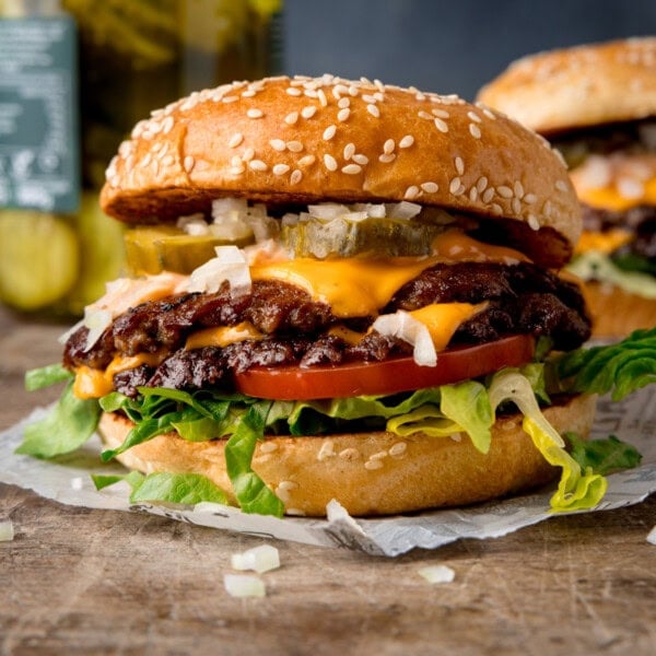 A square image, of the finished smash burgers on a square of white parchment paper. There are a few bits of sliced onion in front of the burger. On the right of the background, there is another burger. On the left side of the background, there is a jar of pickles. This is set on a wooden surface with a blue/grey background.