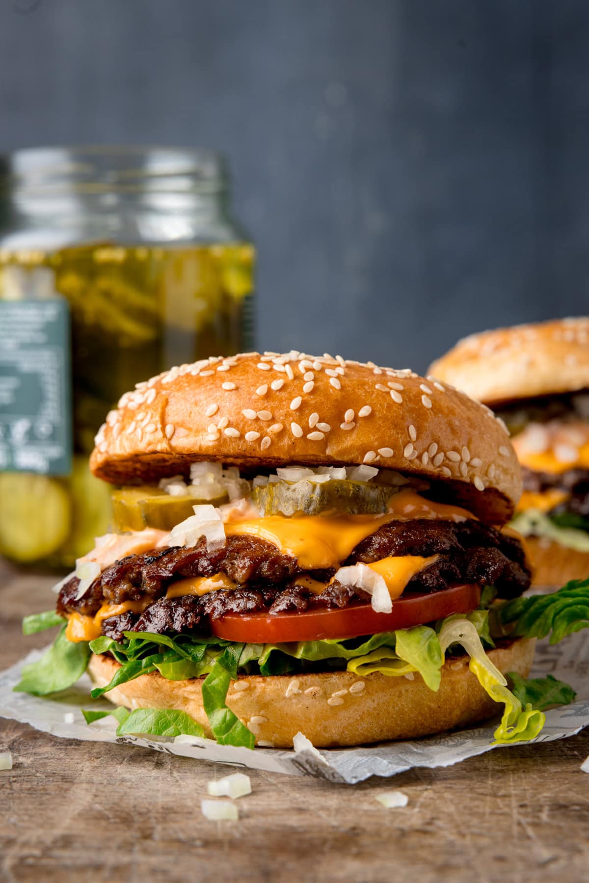 A tall image, of the finished smash burgers on a square of white parchment paper. There are a few bits of sliced onion in front of the burger. On the right of the background, there is another burger. On the left side of the background, there is a jar of pickles. This is set on a wooden surface with a blue/grey background.