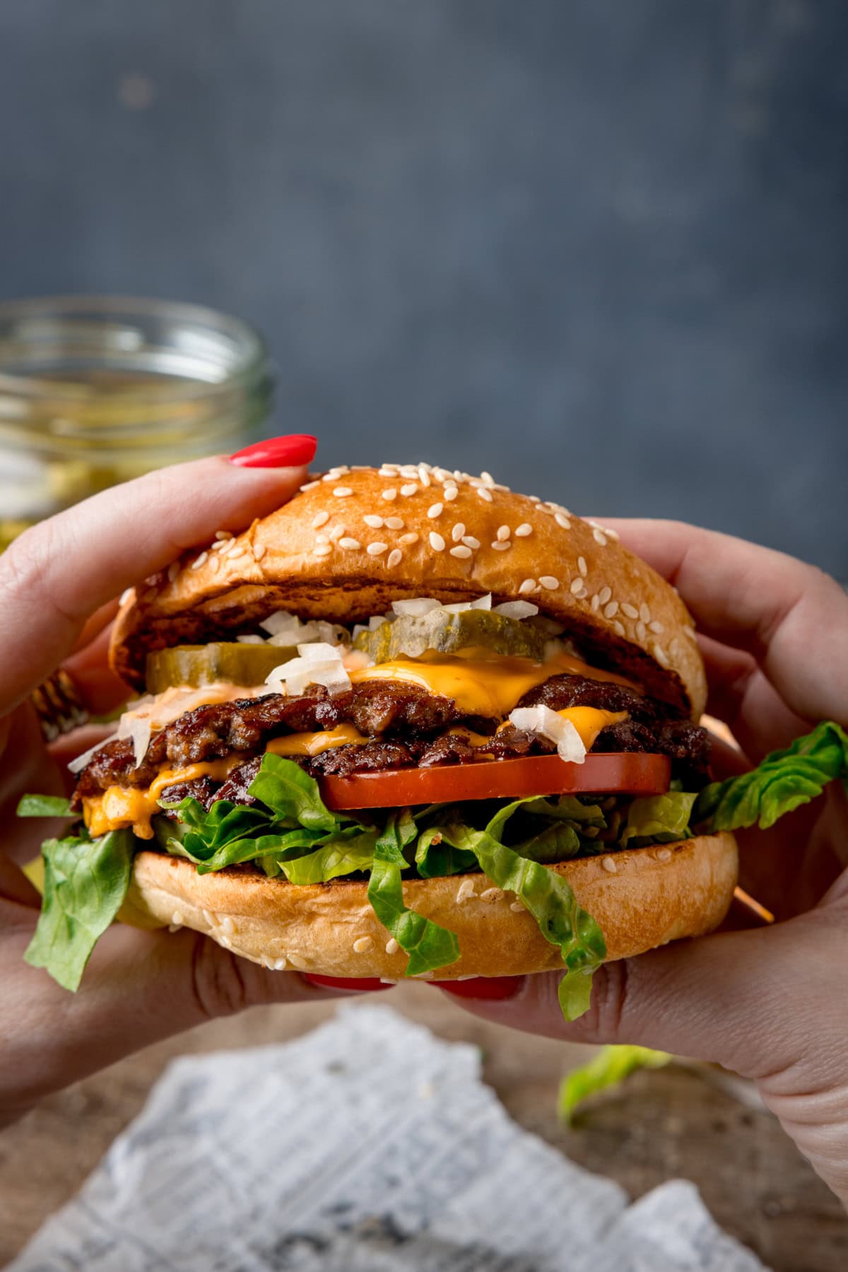 A tall image of the finished smash burgers. In the foreground of the image, there are two hands holding the burger, with the cross-section visible to the screen. In the background, you can see the white parchment paper, a small bit of chopped lettuce (that seems to have fallen from the burger), and a jar of pickles on a wooden surface. This is all set on a blue/grey background.