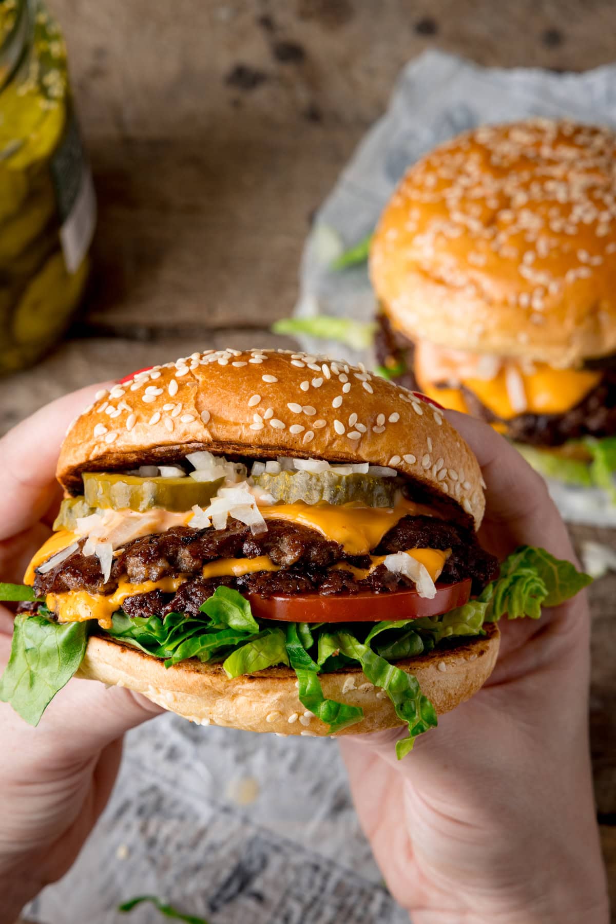 A tall image of the finished smash burgers. In the foreground of the image, there are two hands holding the burger, with the cross-section visible to the screen. In the background, you can see the white parchment paper, a small bit of chopped lettuce (that seems to have fallen from the burger), and, on the left side of the picture, a jar of pickles. On the right side of the background, there is another burger placed on a square of white parchment paper. This is all set on a blue/grey background.