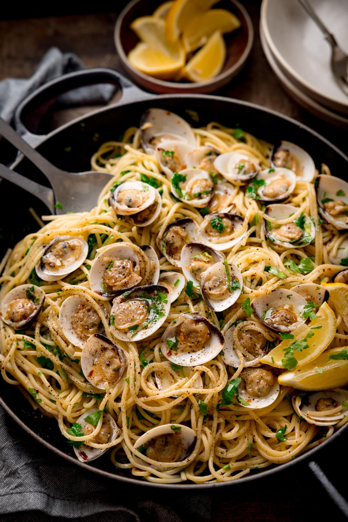 Tall overhead image of Spaghetti Vongole (spaghetti with clams) in a black pan topped with parsley and lemon wedges. There are matt silver serving spoons in the pan. The pan is on a wooden table next to a grey napkin. There are two white bowls with forks and a small bowl of lemon wedges at the top of the frame.