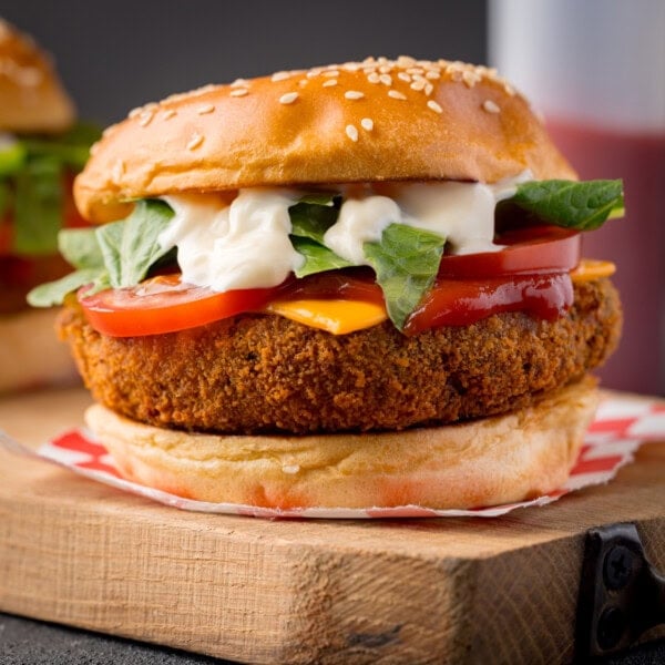 A square, close-up shot of Veggie Bean Burger. The burger is on top of a square of red and white checkered baking parchment. In the left-hand side of the background, you can slightly see a second burger. In the right of the background, you can see a white sauce bottle filled with ketchup. The burger is placed on a wooden cutting board, which is on top of a navy blue surface, This is all set on a navy blue background.