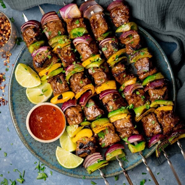 A square overhead shot of steak kebabs. There are five kebabs placed at an angle on a blue plate, beside the skewers, on the left side of the plate there is a small white dish with chilli sauce and four wedges of lime. In the top left of the image, you can slightly see a clear glass jar with dried chilli flakes in it and scattered around it. In the top right of the image, there is a navy blue napkin tucked beneath the main plate. There are dried chilli flakes and shredded coriander scattered around the surface. This is all set on a light blue/grey surface.