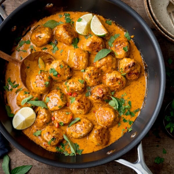 A square, overhead image of Thai Chicken Meatball Curry in a black pan. There is a large wooden spoon on the left side of the dish, scooping some sauce and meatballs. The meatballs are topped with slices of red chilli, and coriander and there are two lime wedges placed in the pan. There is some coriander scattered around the background, and to the left of the pan, there is a dark blue napkin tucked behind the pan. In the top right corner of the image, you can slightly see a stack of light grey dishes with silver cutlery on top. Below the stack of dishes, there is a small black dish filled with chopped coriander. This is all set on a dark grey background.