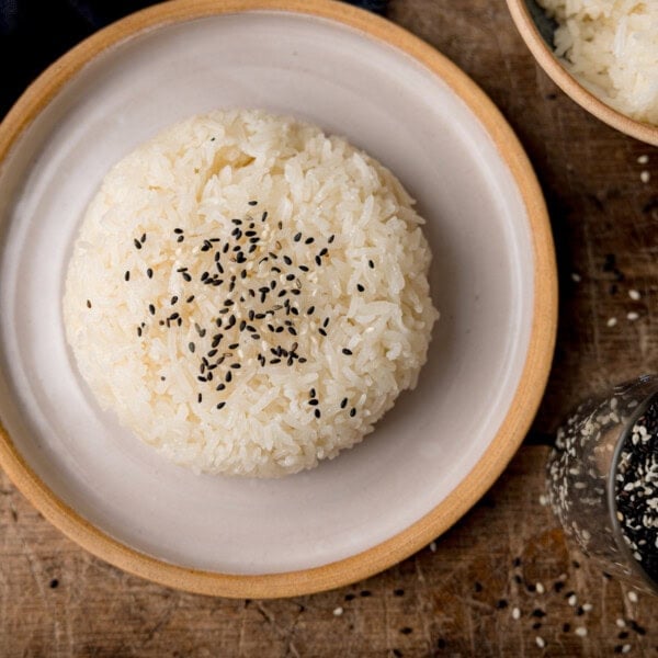 A square, overhead shot of sticky rice on top of a white plate with a light brown lining. The rice is topped with mixed sesame seeds. In the top right corner of the image, you can slightly see the corner of a light brown bowl filled with the rest of the sticky rice. Below that, you can see a clear glass jar filled with mixed sesame seeds, which are also scattered around the jar. This is all set on a dark wooden background.