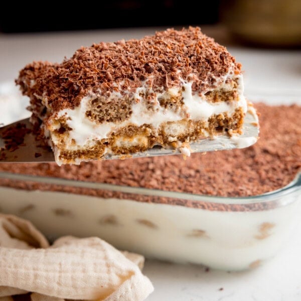 A close up picture of a slice of tiramisu being lifted from a glass dish full of tiramisu.