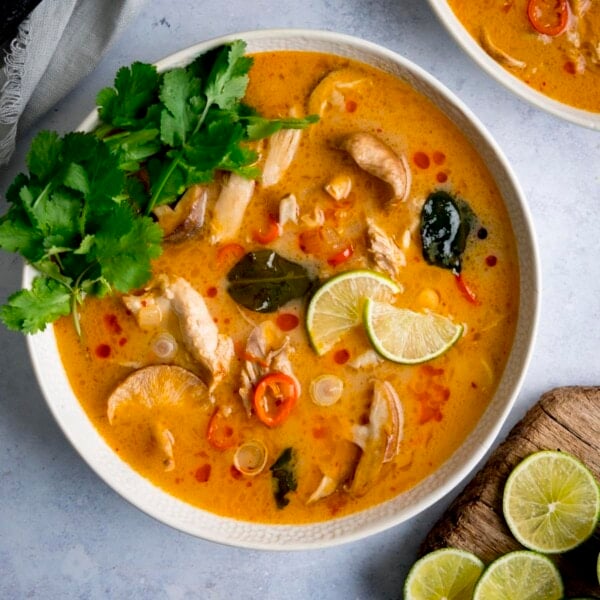 Tom Kha Gai - Thai chicken soup with coconut milk and galangal - in a white bowl on a light background. The soup is garnished with lime slices and coriander.