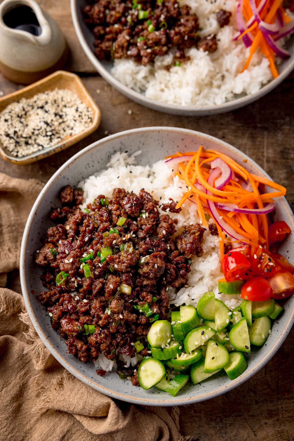 Tall overhead photo of two light blue speckled bowls filled with Vietnamese-style minced beef on a bed of rice, with pickled strips of red onion and carrot. There are also chunks of baby cucumber and chopped baby tomatoes, plus a sprinkling of black and white sesame seeds on each bowl.
The bowls are on a wooden table. There is a small stoneware jug and a small dish of sesame seeds at the top left of the frame.
There is a brown napkin next to the main bowl.