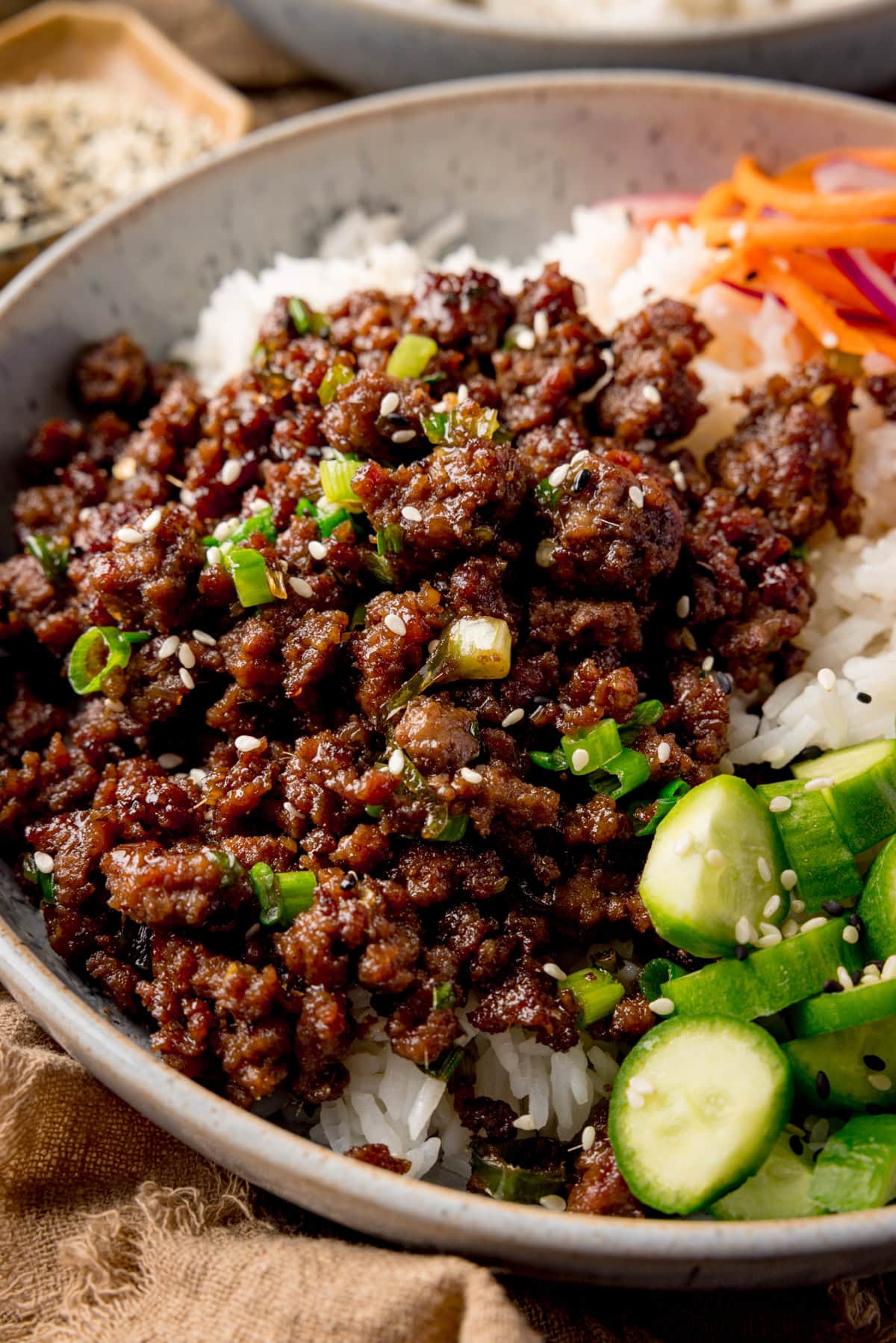 Very close up side-on photo of a light blue speckled bowl filled with Vietnamese-style minced beef on a bed of rice, with pickled strips of red onion and carrot. There are also chunks of baby cucumber and a sprinkling of black and white sesame seeds in the dish. 
There is a small dish of sesame seeds at the top left of the frame.
There is a brown napkin next to the bowl at the bottom left of the frame.