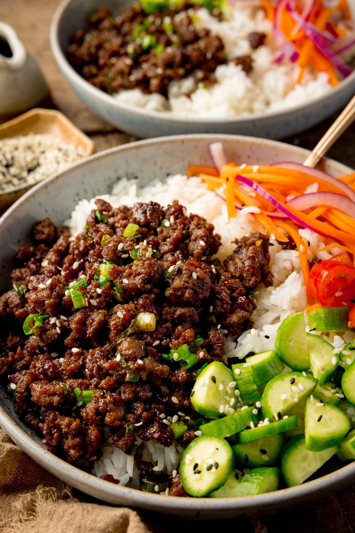 Close up side-on photo of a light blue speckled bowl filled with Vietnamese-style minced beef on a bed of rice, with pickled strips of red onion and carrot. There are also chunks of baby cucumber and chopped baby tomatoes, plus a sprinkling of black and white sesame seeds. There is a further, similar bowl in the background.
The bowls are on a wooden table. There is a small stoneware jug and a small dish of sesame seeds at the top left of the frame.
There is a brown napkin next to the main bowl.