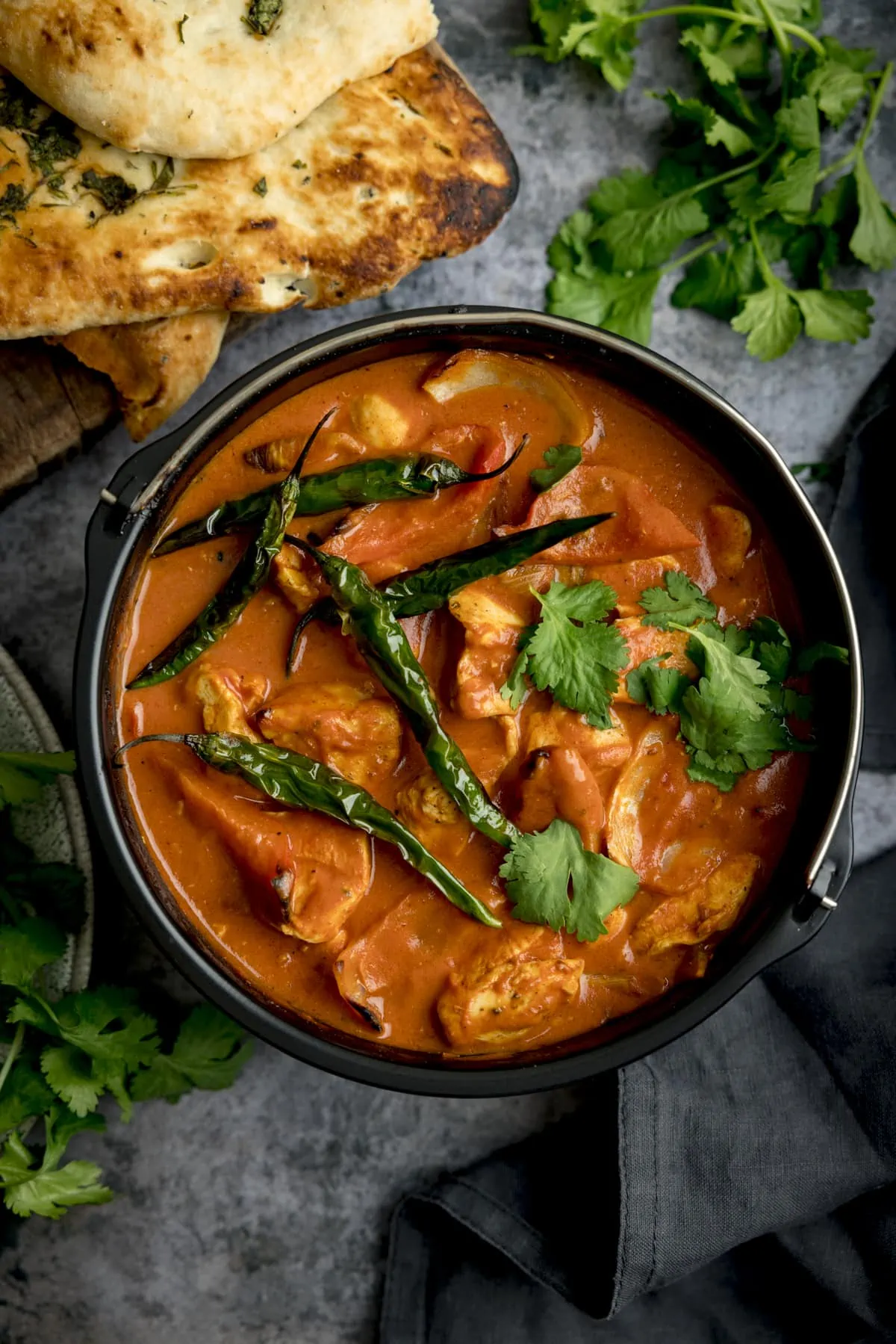 A Tall overhead shot of Air Fryer Chicken Curry sprinkled with coriander and whole fried green chillies, with bunches of coriander scattered around, there is a pile of toasted naan breads on a wooden board, some dark fabric napkins are to the side, against a mottle grey work surface.