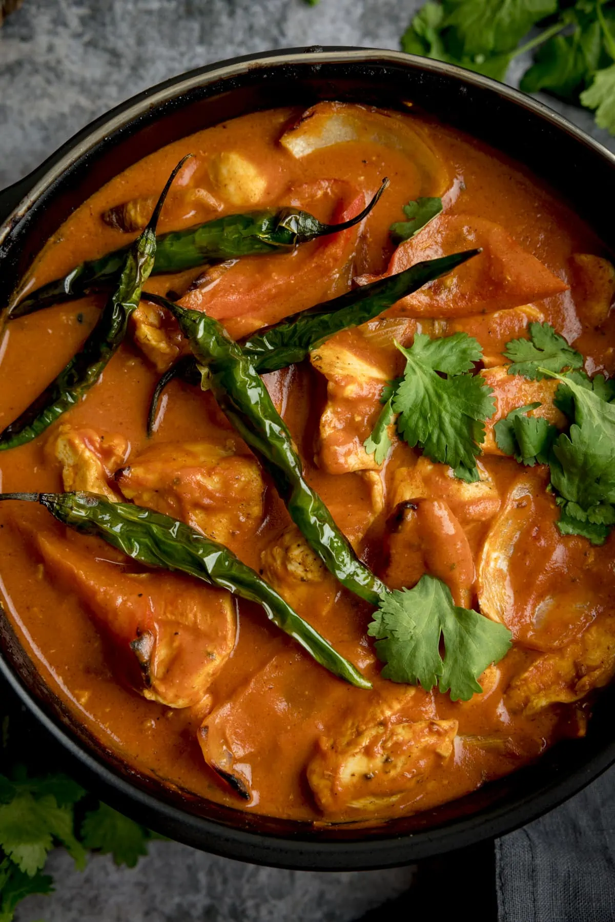 Close up overhead shot of Air Fryer Chicken Curry sprinkled with coriander and whole fried green chillies, with bunches of coriander scattered around, some dark fabric napkins are to the side, against a mottle grey work surface.