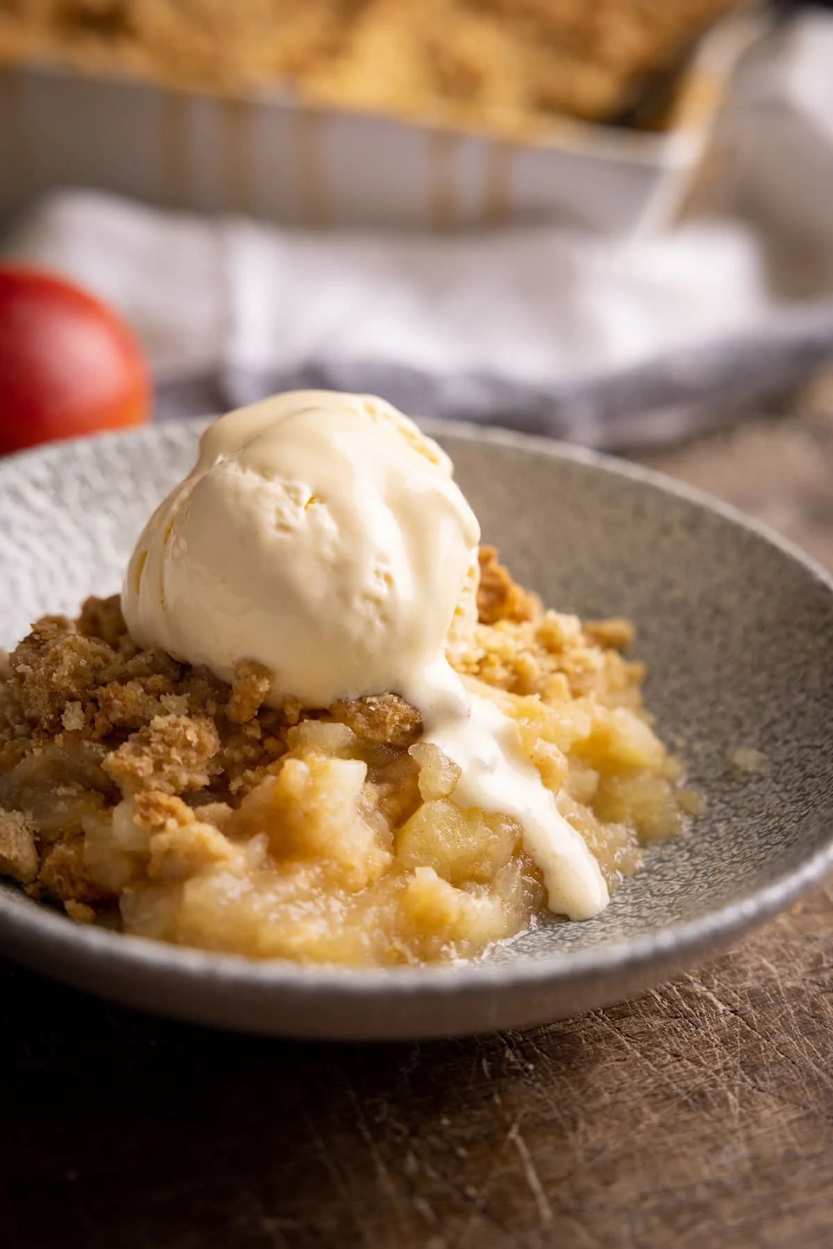 Close up photo of a bowl with a portion of apple crumble in and a blob of ice cream slowly melting onto the hot crumble.