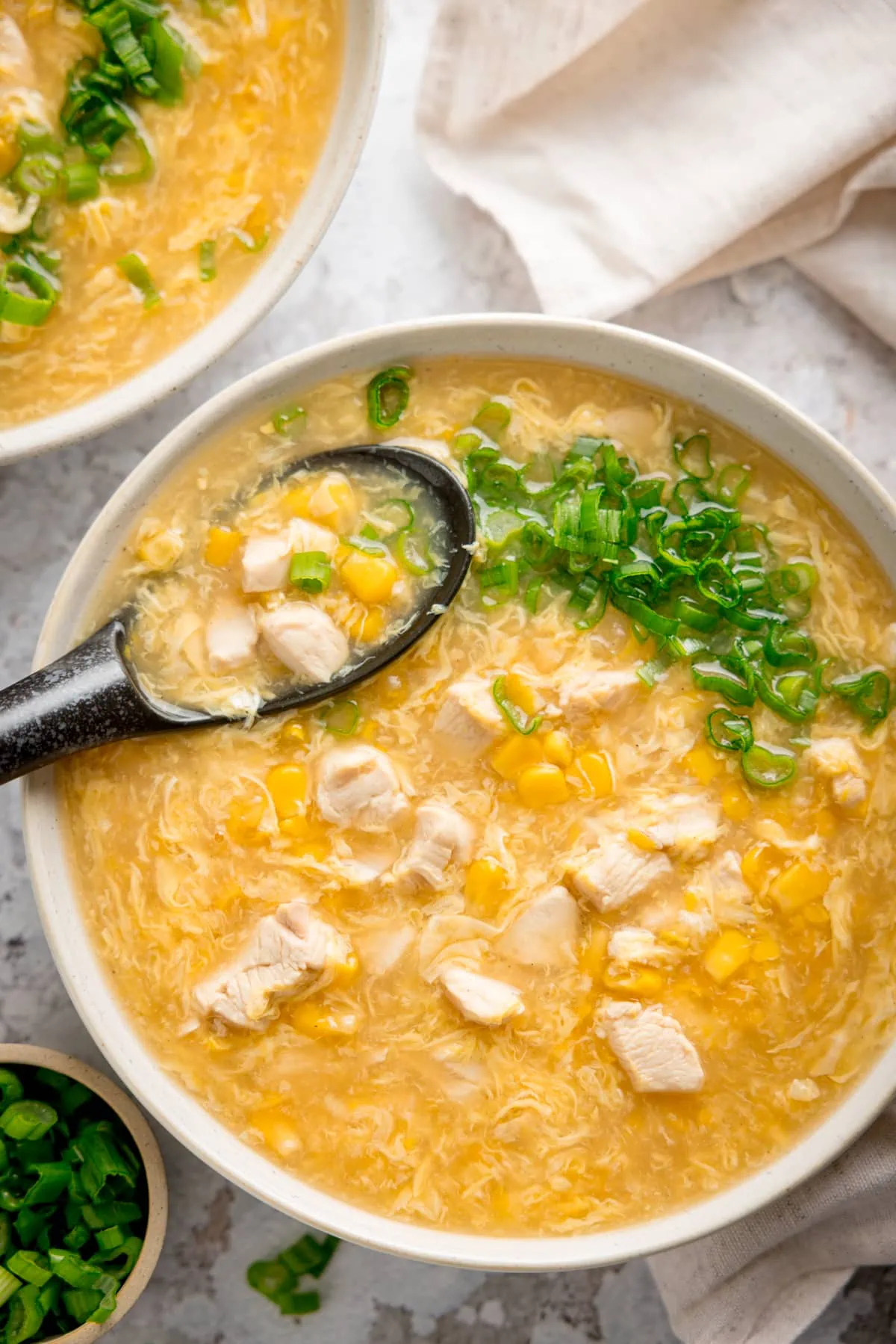 Bowl of chicken and sweetcorn soup with spring onions on top and a black spoon sticking out. There is a further bowl of soup in the top of the shot, and small bowl of spring onions in the bottom of the shot and a cream napkin next to the bowls of soup.