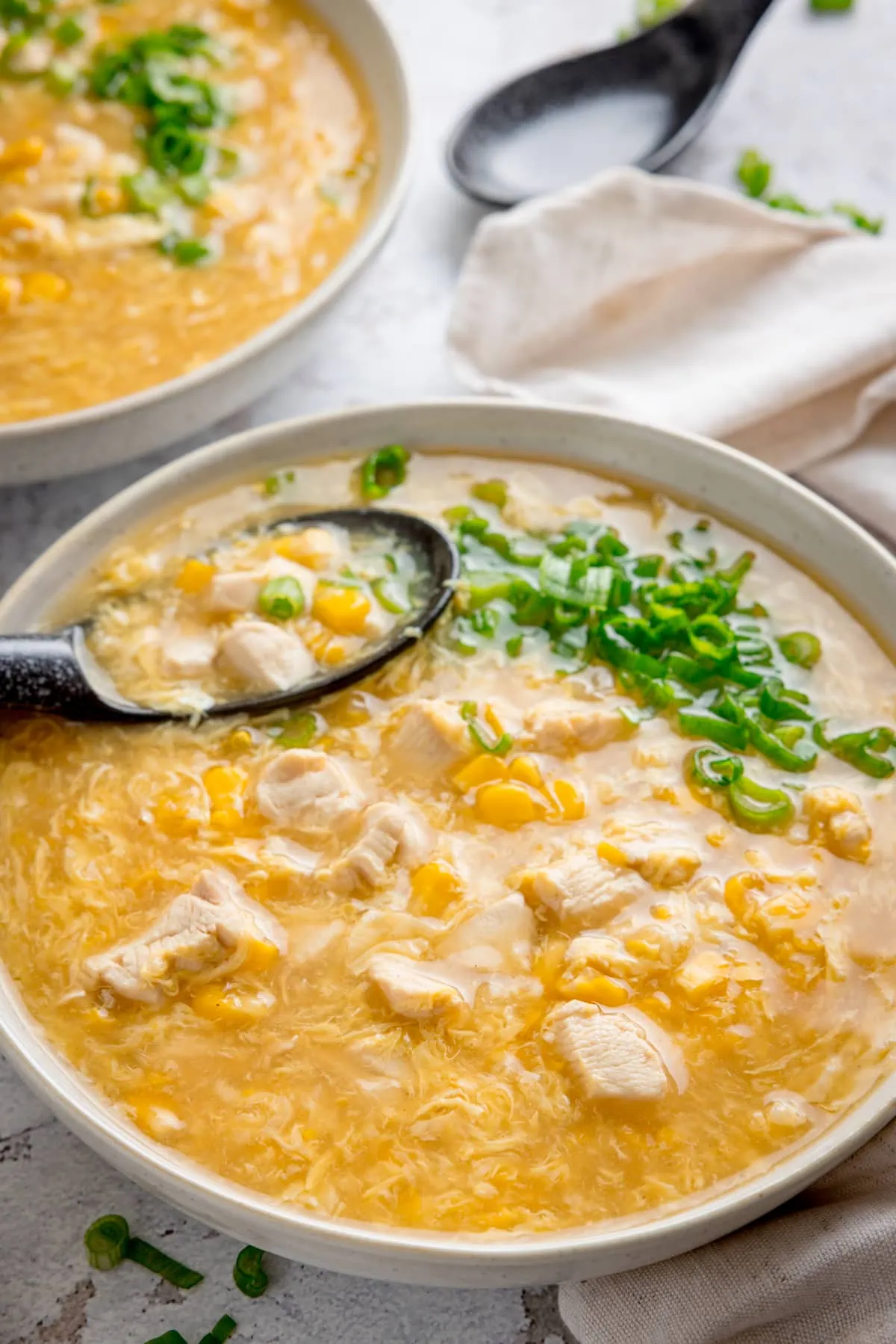 Bowl of chicken and sweetcorn soup with spring onions on top and a black spoon sticking out. There is a further bowl of soup in the background, along with a another black spoon and a cream napkin.