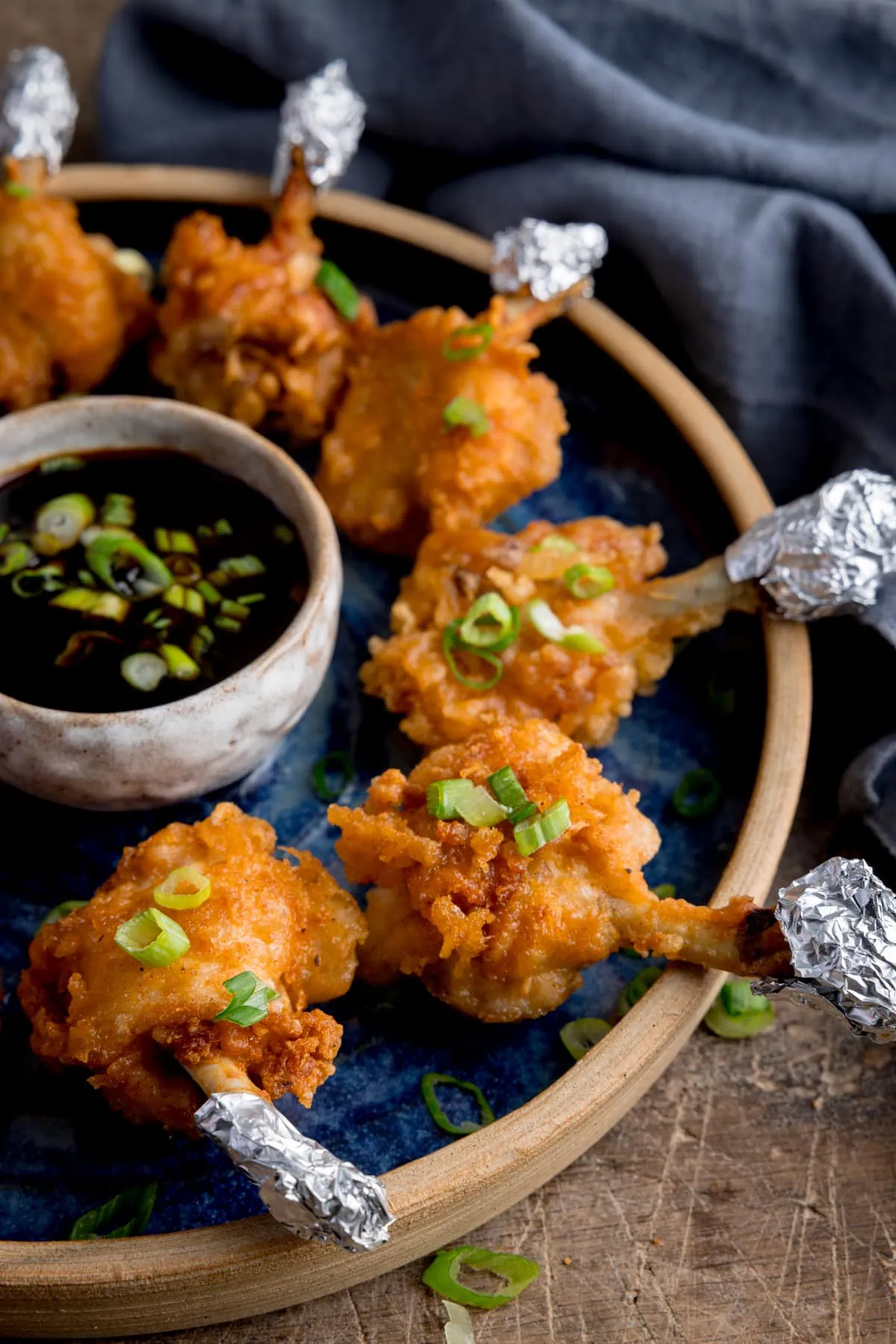 Close up side image of chicken lollipops arranged in a circle on a blue plate with a dipping bowl of sticky spicy sauce int he middle. The lollipops and sauce have been sprinkled with chopped spring onions and the plate is in a wooden table next to a blue napkin.