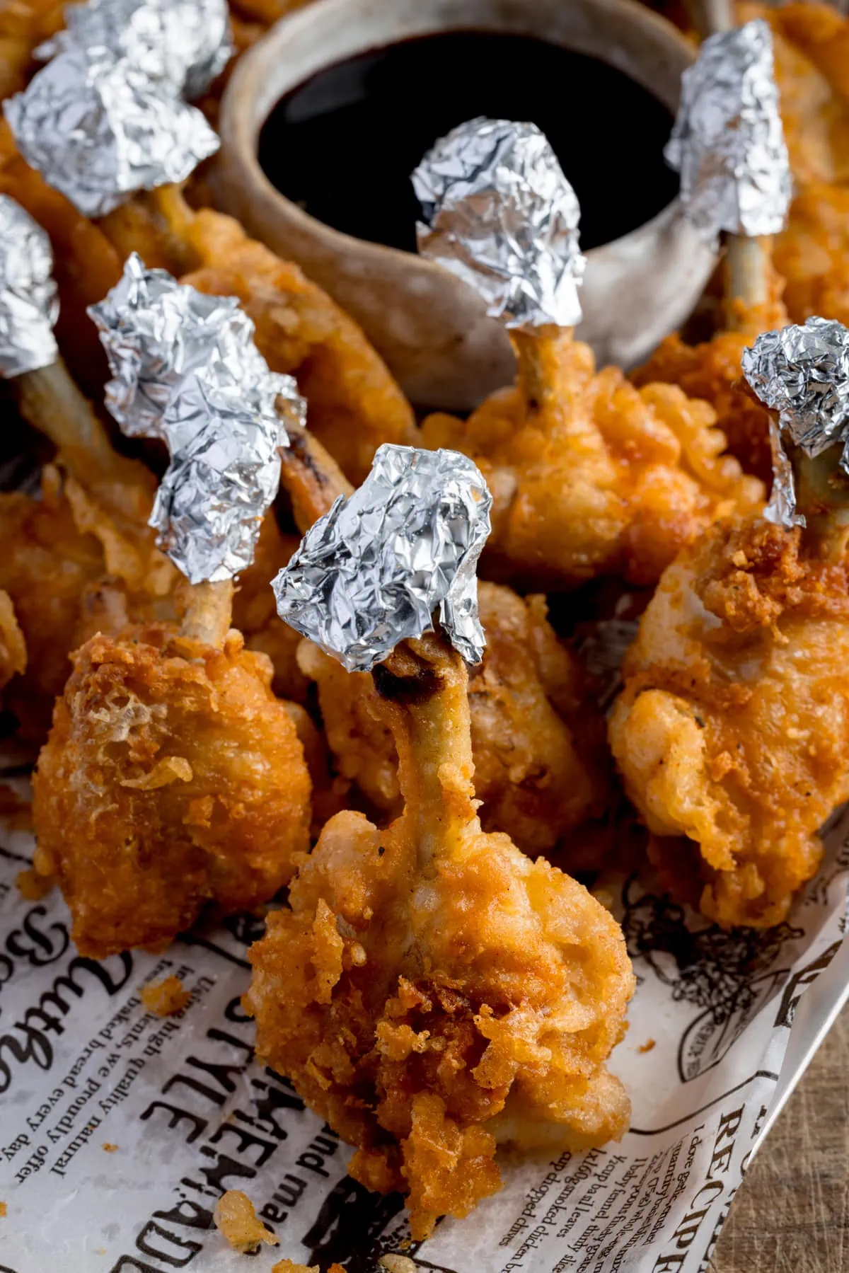 Chicken lollipops, standing up on a piece of white baking paper that's decorated with newspaper-style writing. The lollipops have been wrapped in foil at the ends and they're next to a small light bowl of a dark dipping sauce.