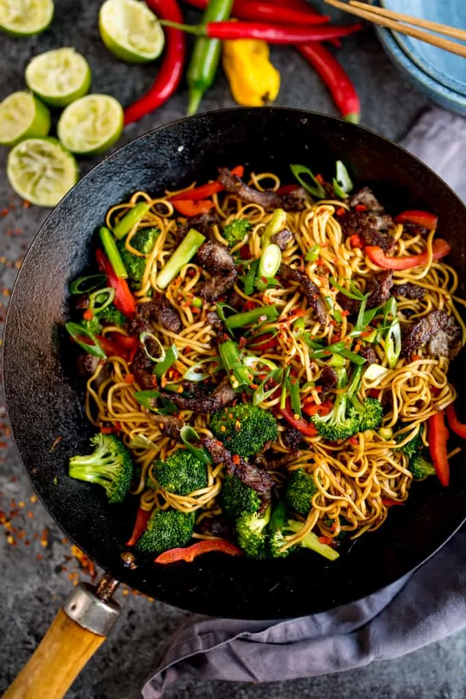 Overhead image of crispy chilli beef noodles with red pepper and broccoli in a wok.
The wok is on a grey surface and there are ingredients scattered around. There is also a grey napkin, blue bowls and a set of chopsticks in shot.